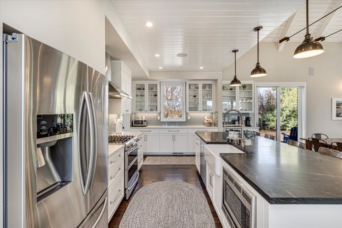 Bright modern farmhouse kitchen — sunlit open-concept space with a long dark stone island, farmhouse sink, stainless steel French-door refrigerator, gas range, white cabinets with glass-front uppers, pendant lights, and sliding glass door to the backyard.