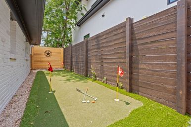 Narrow residential side-yard backyard putting green with artificial turf, three practice flags and putters, brown wooden privacy fence, white brick house wall and decorative wooden gate with star