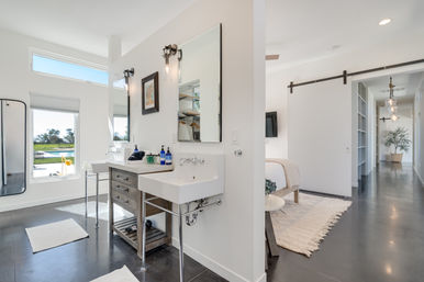 Bright modern bathroom in a sunlit home with dual sinks, wood vanity, large mirrors, a barn-style sliding door to the bedroom, and window views of a backyard pool and lawn.
