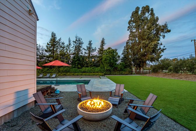 Cozy backyard patio at dusk with a round stone fire pit surrounded by Adirondack chairs, adjacent swimming pool with red umbrellas, wide green lawn and tall trees