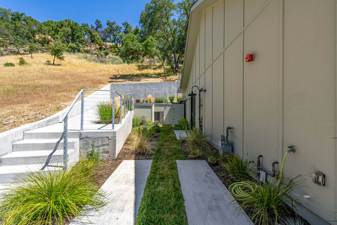 Sunny hillside exterior walkway beside a beige modern building with concrete steps and path, metal cable railing, narrow grass strip, drought-tolerant grasses, utility hookups, and oak trees in the background.