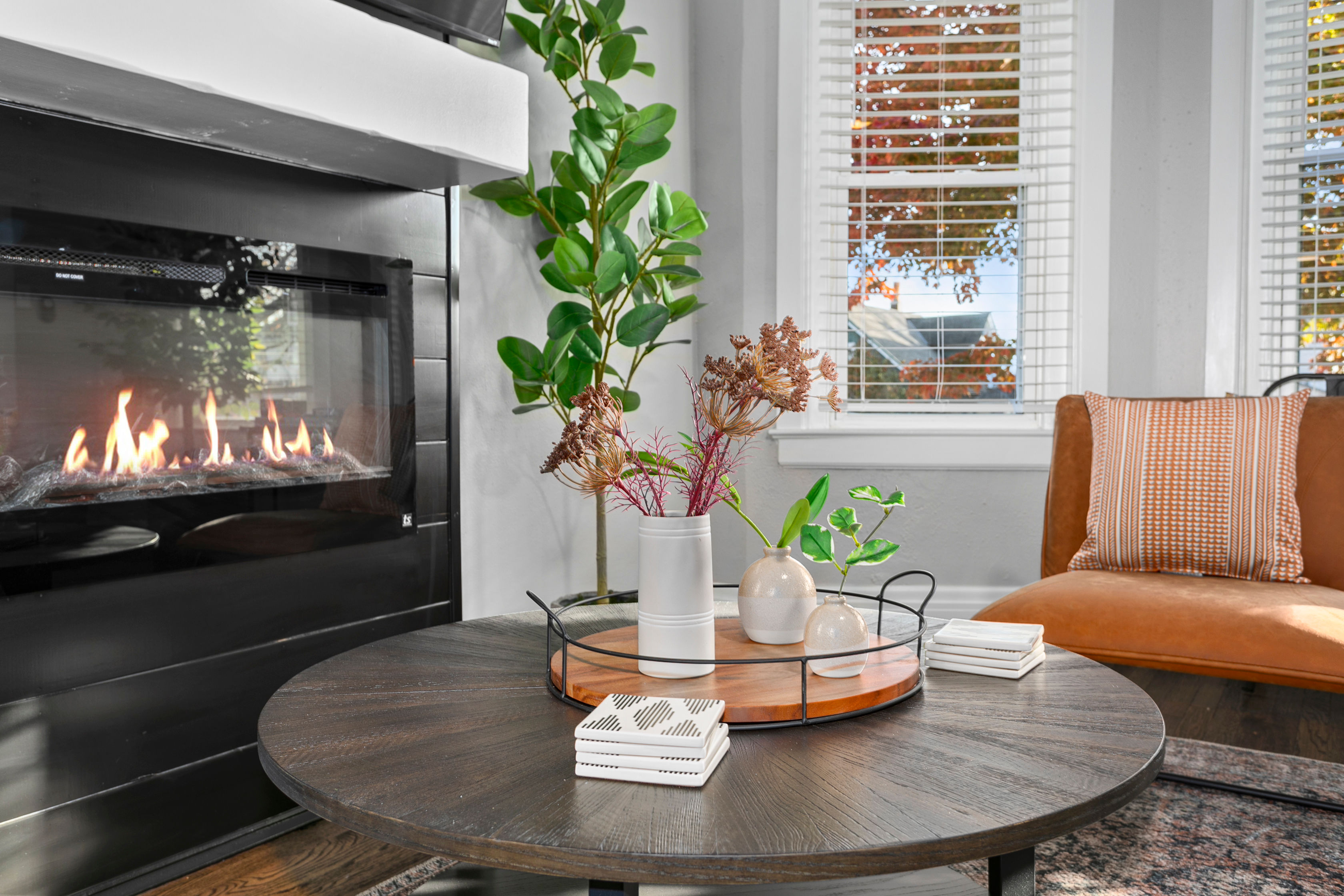 Cozy modern living room with a lit gas fireplace, round dark-wood coffee table holding a tray of vases and greenery, a tan leather accent chair, and a window with autumn foliage outside.