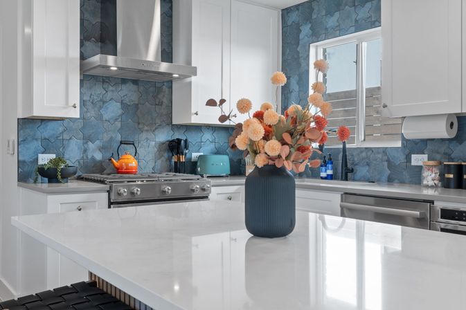 Bright contemporary kitchen with white cabinets and quartz island, blue geometric tile backsplash, stainless steel range and hood, teal toaster and orange kettle, and a ribbed navy vase of peach and coral flowers as a colorful centerpiece.