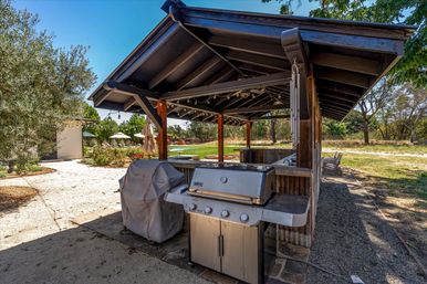 Sunny outdoor kitchen and backyard pavilion with a stainless steel gas grill, covered wooden pergola with string lights, patio seating, umbrellas and lawn garden