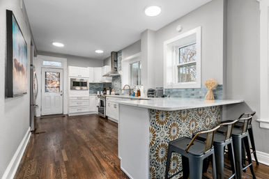 Bright modern kitchen with white cabinets, stainless steel appliances, hardwood floors, peninsula island with colorful geometric tile facing and three metal bar stools, blue glass-tile backsplash and windows