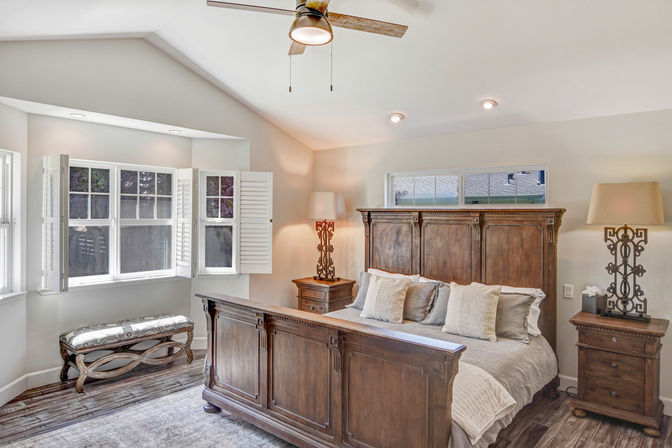 Cozy sunlit master bedroom with a large rustic wooden bed and matching nightstands, neutral bedding and pillows, decorative lamps, bay window with white plantation shutters and upholstered bench, ceiling fan and hardwood floors.