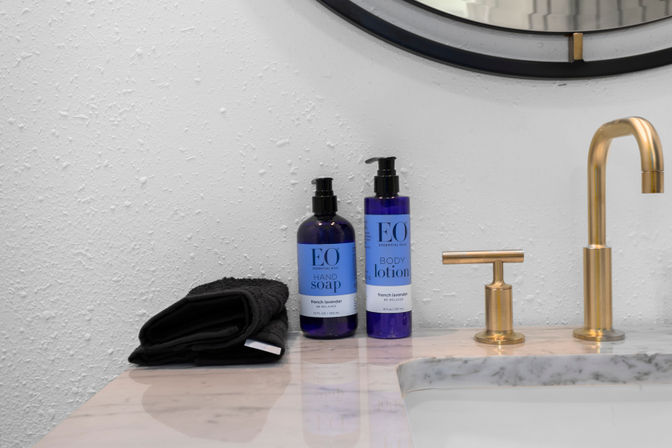 Modern bathroom vanity with marble countertop, gold faucet, black folded towel and blue pump bottles of lavender hand soap and body lotion beneath a round mirror.
