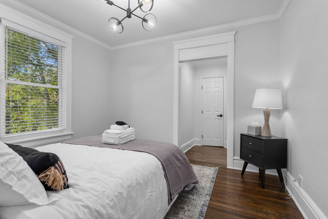 Bright staged bedroom with white walls and hardwood floors, white bed topped with a mauve throw and folded towels, black mid-century nightstand with lamp, window blinds showing green trees, and a modern ceiling light fixture.