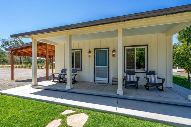 Inviting sunny front porch of a modern farmhouse with white board-and-batten siding, teal entry door and lanterns, four black rocking chairs with striped cushions on a covered concrete stoop, green lawn and adjacent wooden carport under a clear blue sky.