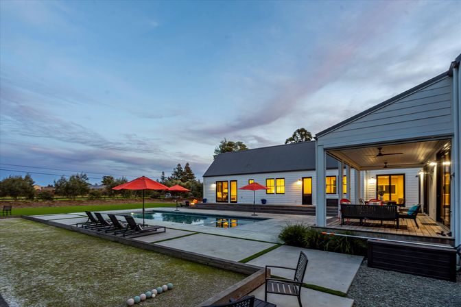 Modern farmhouse at dusk — lit interior windows, covered porch with dining seating and ceiling fans, rectangular swimming pool with loungers and red umbrellas, landscaped lawn and bocce balls under a wide evening sky.