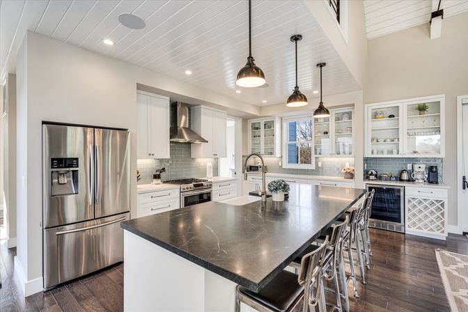 Bright modern farmhouse kitchen with large dark stone island, stainless-steel French-door fridge, gas range, white shaker cabinets, glass-front uppers, pendant lights and chrome bar stools in an open-concept home