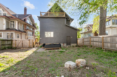 Sunny urban backyard with a gray three-story house and rooftop deck, fenced yard with a small patio and outdoor sectional, large shade tree and neighboring vintage homes