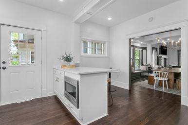 Bright open-concept kitchen with white marble island and built-in microwave, dark hardwood floors, glass-paned entry door, and adjoining cozy dining and living area with bench seating and chandelier.