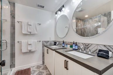 Spa-like modern bathroom with double sink vanity and dark countertops, round mirrors, patterned tile floor, glass-enclosed shower and neatly folded white towels.