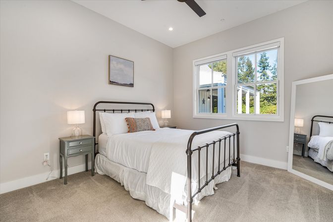 Bright sunlit bedroom with black iron bed and crisp white linens, patterned throw pillow, matching nightstands with lamps, beige carpet, large window with tree view and a leaning full-length mirror.