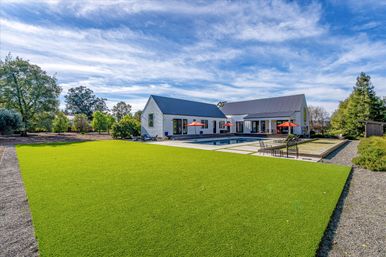 Sunlit modern farmhouse backyard with metal-roof house, large rectangular pool, bright green lawn, patio seating and orange umbrellas under a blue sky.