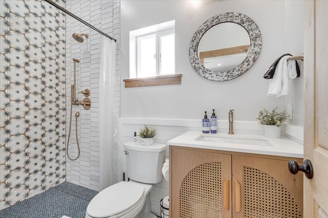 Bright modern farmhouse bathroom with starburst-pattern shower tile, brass shower fixtures, blue mosaic floor, wood rattan vanity with white countertop, round mirror, toilet and small window