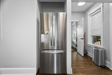 Gleaming stainless-steel French-door refrigerator with bottom freezer and water dispenser set into a white-walled kitchen hallway with hardwood floors, white trim, trash bin and cleaning tools visible.