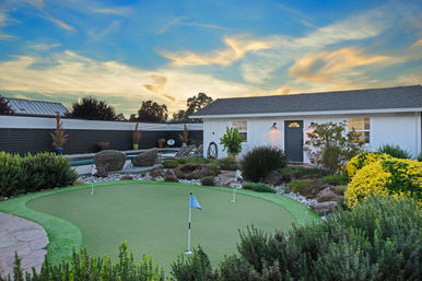 Sunset suburban backyard with a small artificial putting green and three flags, modern pool with wicker loungers, drought-tolerant landscaping, and a white single-story cottage under a colorful streaked sky.