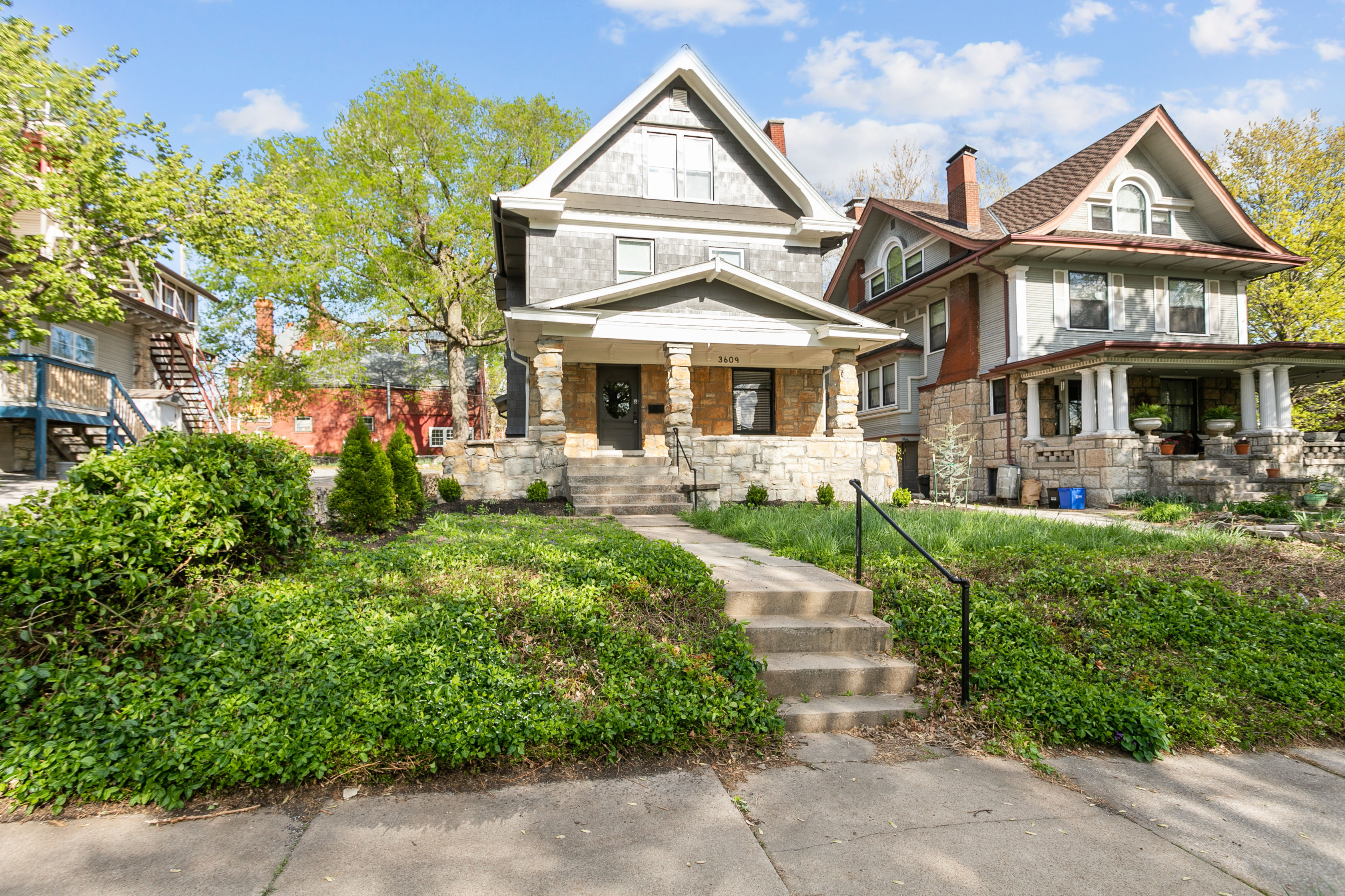 Cheerful historic Craftsman-style house with a stone front porch and concrete steps on a tree-lined residential street, flanked by neighboring early-1900s homes under a bright blue sky.