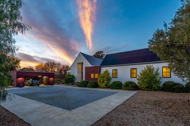 Modern farmhouse-style home with rooftop solar panels, warm lit windows, detached two-car garage with SUV, gravel driveway and manicured shrubs under a dramatic pink-orange sunset sky.