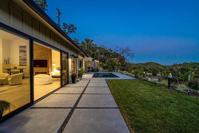 Modern hillside home at twilight with floor-to-ceiling sliding glass doors revealing a cozy, warmly lit living room; concrete slab patio leads past a manicured lawn to a rectangular plunge pool and rolling hill views.
