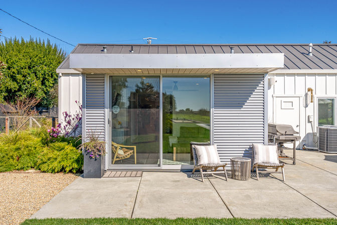 Modern backyard patio with sliding glass doors opening into a small white studio, two wicker lounge chairs with striped cushions and a round side table on a concrete patio, potted plants, barbecue grill and green lawn under a clear blue sky.