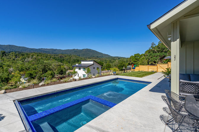 Sunlit modern backyard pool with built-in spa and concrete patio, wicker seating at right, grassy yard and wooden fence beyond, sweeping forested hills and clear blue sky