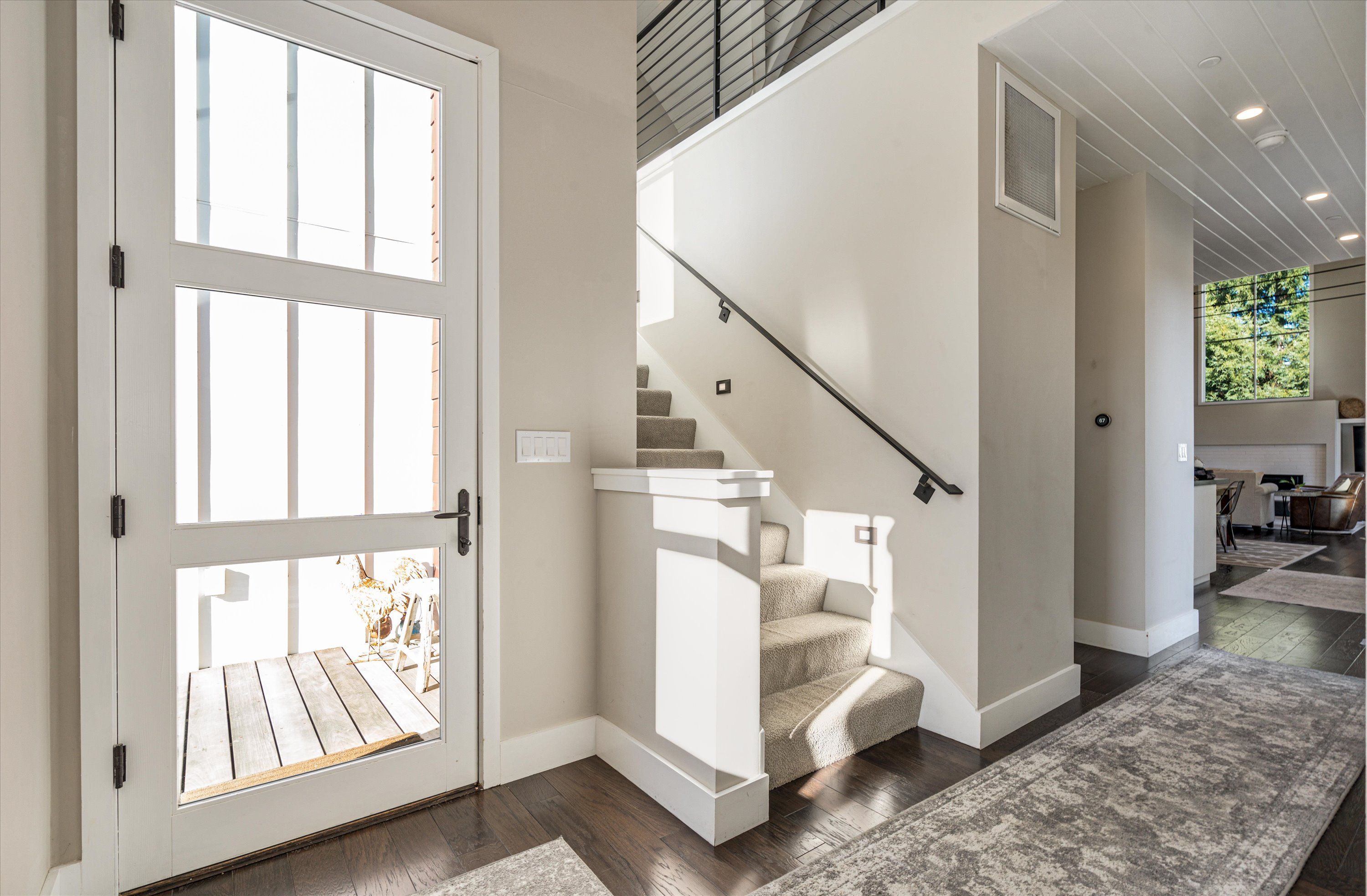 Sunlit modern foyer in a contemporary home with a glass‑paned front door, carpeted stairs and black metal handrail, dark hardwood floors and a view into the open living area.