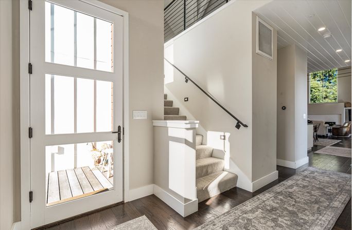 Sunlit modern foyer in a contemporary home with a glass‑paned front door, carpeted stairs and black metal handrail, dark hardwood floors and a view into the open living area.