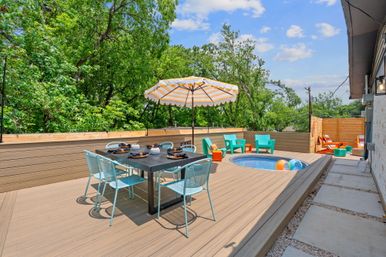 Sunny backyard deck patio with round built-in plunge pool, black dining table with turquoise chairs, striped umbrella and colorful lounge seating amid green trees