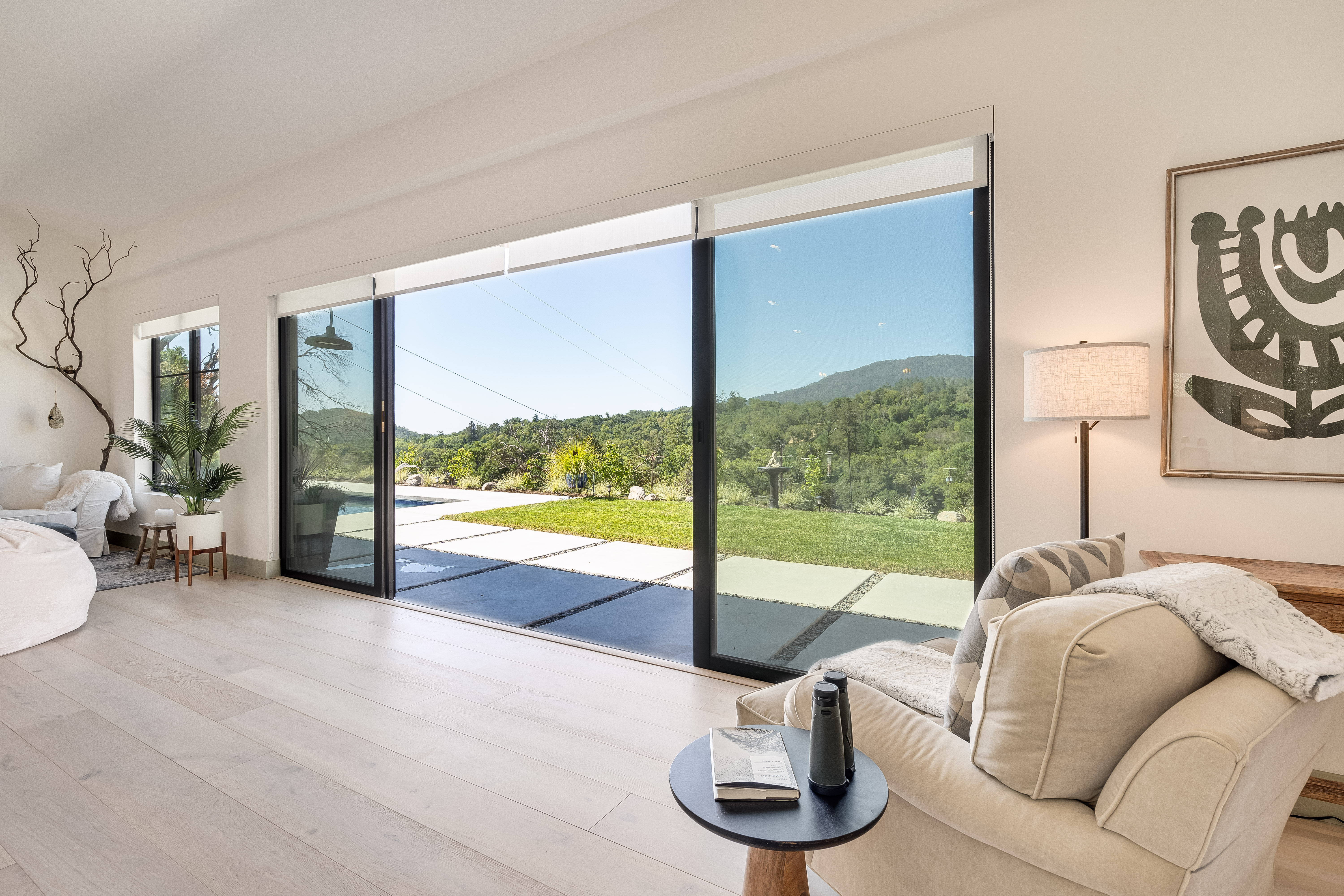 Sunlit modern living room with floor-to-ceiling sliding glass doors opening to a patio and pool, manicured lawn and rolling green hills under a clear blue sky; neutral armchair, side table with water bottle and notebook, potted plants and minimalist wall art.