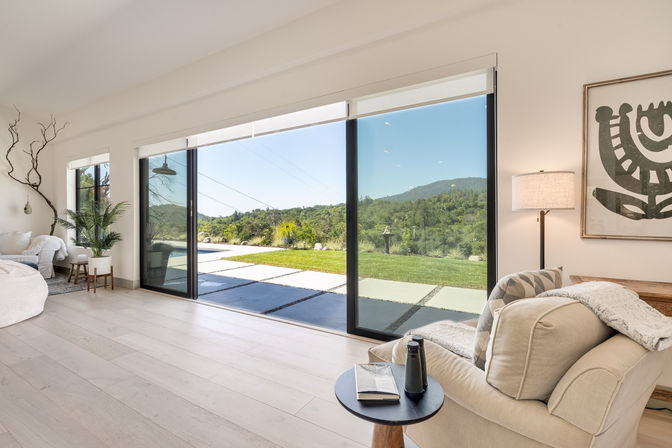 Sunlit modern living room with floor-to-ceiling sliding glass doors opening to a patio and pool, manicured lawn and rolling green hills under a clear blue sky; neutral armchair, side table with water bottle and notebook, potted plants and minimalist wall art.