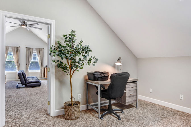 Cozy attic loft home office with sloped ceiling: wooden desk with printer and task lamp, black swivel chair, woven-basket potted plant, and open doorway to a bright sitting area with windows and curtains.