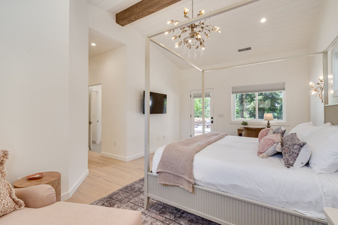 Bright modern bedroom with a four-poster bed, white linens and blush throw, crystal chandelier and exposed wood beam, chaise lounge, wall TV and French doors to a garden