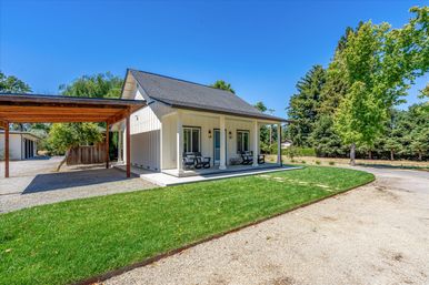 Bright modern farmhouse-style cottage with a covered front porch and rocking chairs, wooden carport, manicured green lawn and gravel driveway under a clear blue sky surrounded by mature trees.