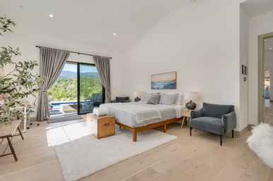 Sunlit modern master bedroom with wooden platform bed, plush white rug, gray armchairs and nightstands, sliding glass doors opening to a patio and pool with lush mountain views.