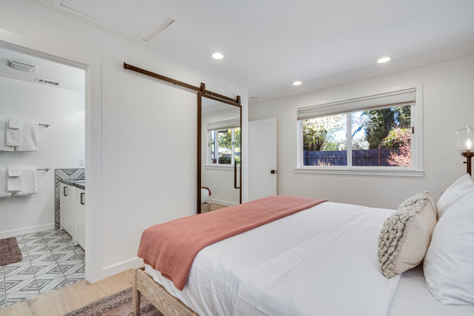 Sunlit modern bedroom with white walls, wooden bed in white linens and coral throw, cozy textured pillow, sliding barn-style mirror door to en-suite bathroom and large window with backyard view