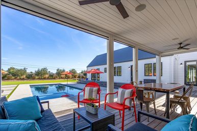 Covered backyard patio overlooking a rectangular pool in a sunny suburban yard, with red metal lounge chairs and umbrellas, blue cushioned seating, a wooden dining table, and a white farmhouse-style house.