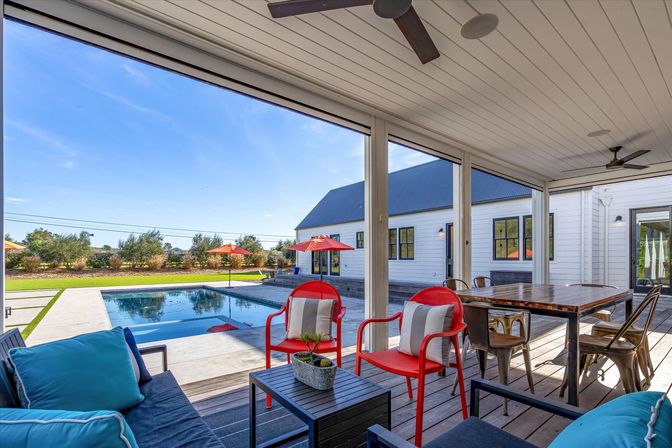 Covered backyard patio overlooking a rectangular pool in a sunny suburban yard, with red metal lounge chairs and umbrellas, blue cushioned seating, a wooden dining table, and a white farmhouse-style house.