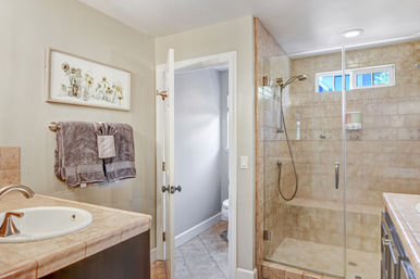 Bright residential bathroom with glass-enclosed beige tiled walk-in shower, single sink vanity with stone countertop, gray towels on a rack, and framed floral wall art.