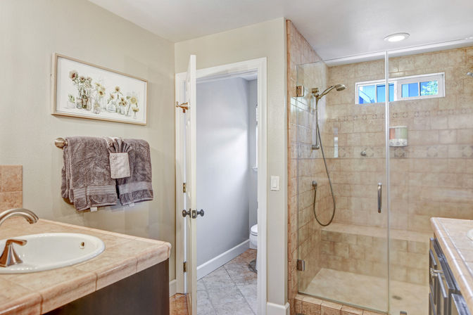 Bright residential bathroom with glass-enclosed beige tiled walk-in shower, single sink vanity with stone countertop, gray towels on a rack, and framed floral wall art.