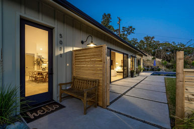 Dusk exterior of a modern single-story home with warm interior lighting visible through sliding glass doors, concrete paver walkway, wooden privacy screen and bench, and a backyard pool near a hillside