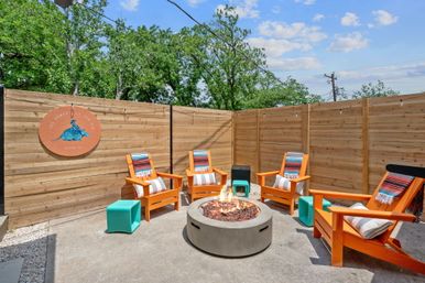 Sunlit Austin, TX fenced patio with a round concrete fire pit, five bright orange Adirondack chairs draped with colorful striped blankets, turquoise stools, wooden privacy fence and trees under a blue sky.