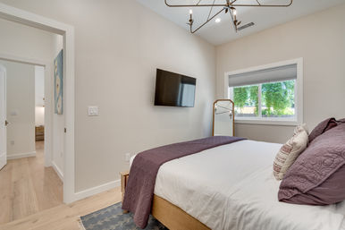 Cozy modern bedroom interior with light wood floors, white bedding and purple quilt, wall-mounted flat-screen TV, full-length mirror by a window overlooking leafy backyard, minimalist decor and natural light.
