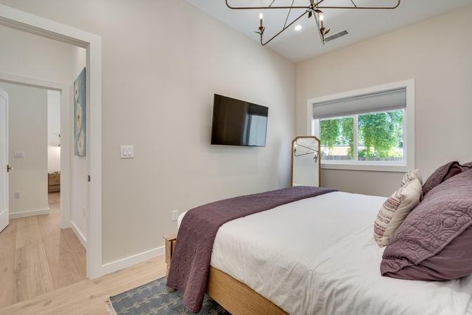 Cozy modern bedroom interior with light wood floors, white bedding and purple quilt, wall-mounted flat-screen TV, full-length mirror by a window overlooking leafy backyard, minimalist decor and natural light.