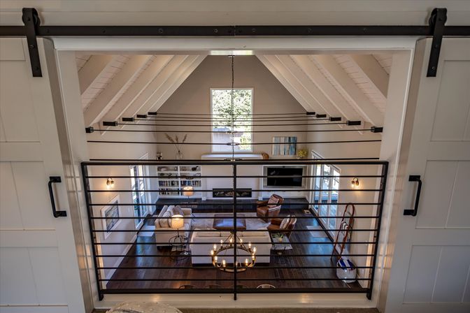 Loft view of an open-concept living room with vaulted white-beamed ceiling, sliding barn doors, black metal railing, chandelier, hardwood floors, neutral sofas and built-in shelving
