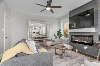 Contemporary open-concept living room with a gray sofa and yellow throw, round wood coffee table, cozy armchair, wall-mounted TV above a linear fireplace, ceiling fan, large window, and a view into the dining area and white kitchen.