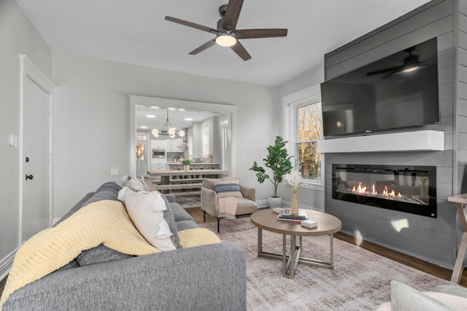 Contemporary open-concept living room with a gray sofa and yellow throw, round wood coffee table, cozy armchair, wall-mounted TV above a linear fireplace, ceiling fan, large window, and a view into the dining area and white kitchen.