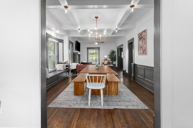 Sunlit open-concept dining and living room in a craftsman-style home with a long wooden farmhouse table, benches and a white spindle chair on a patterned rug, coffered ceiling with brass chandelier, gray wainscoting and hardwood floors, built-in window bench and sofa by large windows with leafy outdoor view.